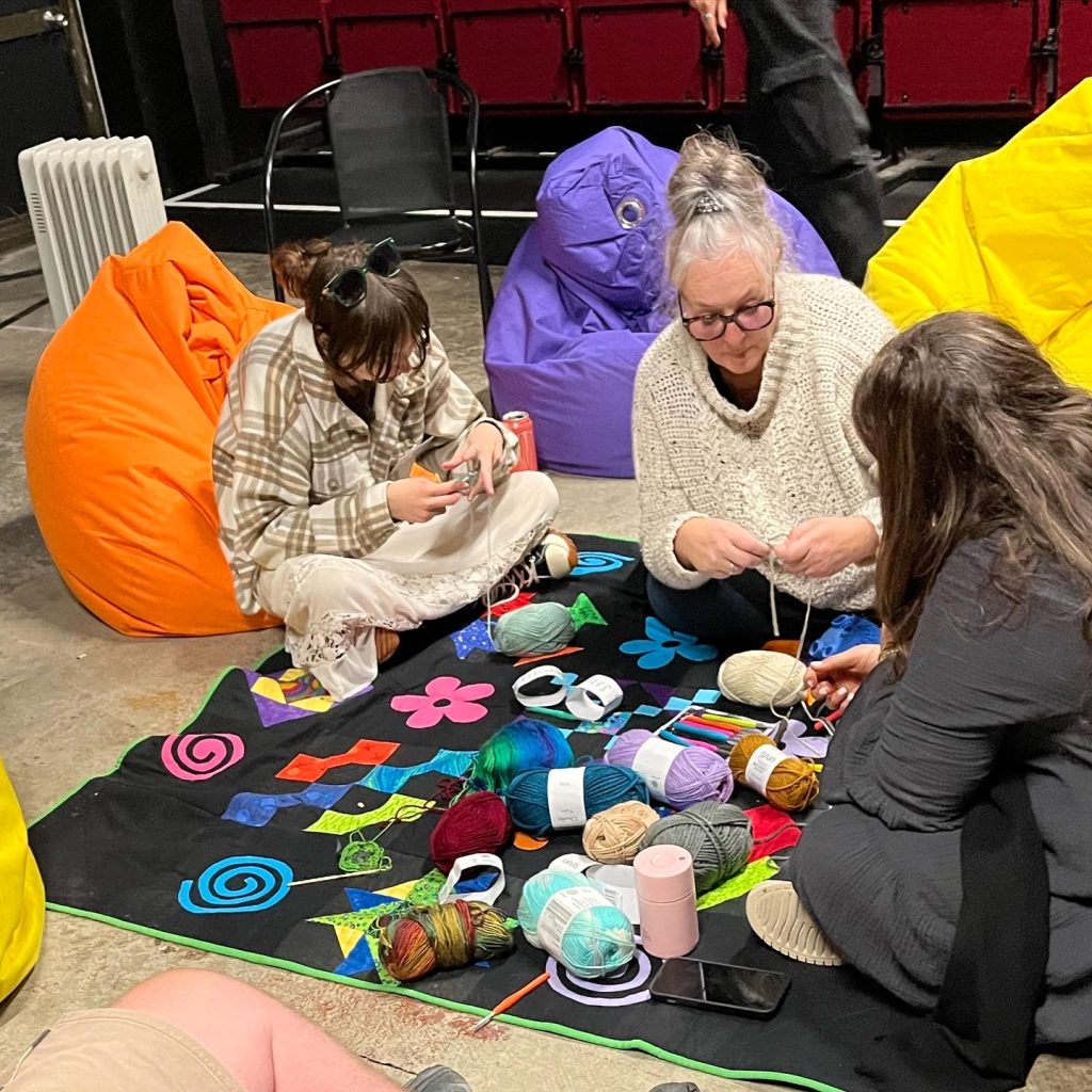 Three women sitting on a colourful patchwork rug doing crochet with lots of balls of wool and orange, purple and yellow beanbags in the background
