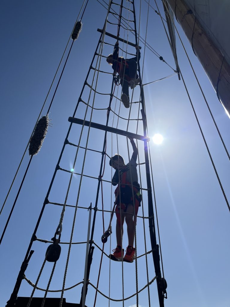 Two youth climbing the mast rigging silhouetted by the sun against bright blue sky