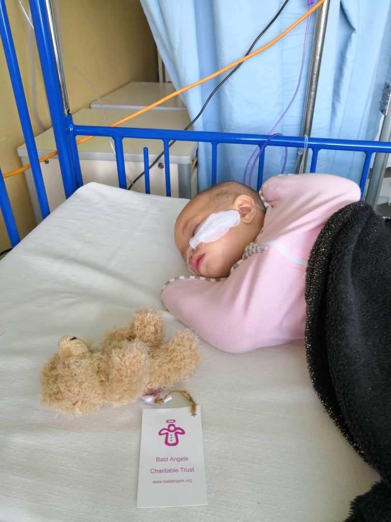 A very small baby in pink with a bandage on her cheek and tube in nose, lies on a hospital bed with bright blue frame. Beside her is an Angel Bear