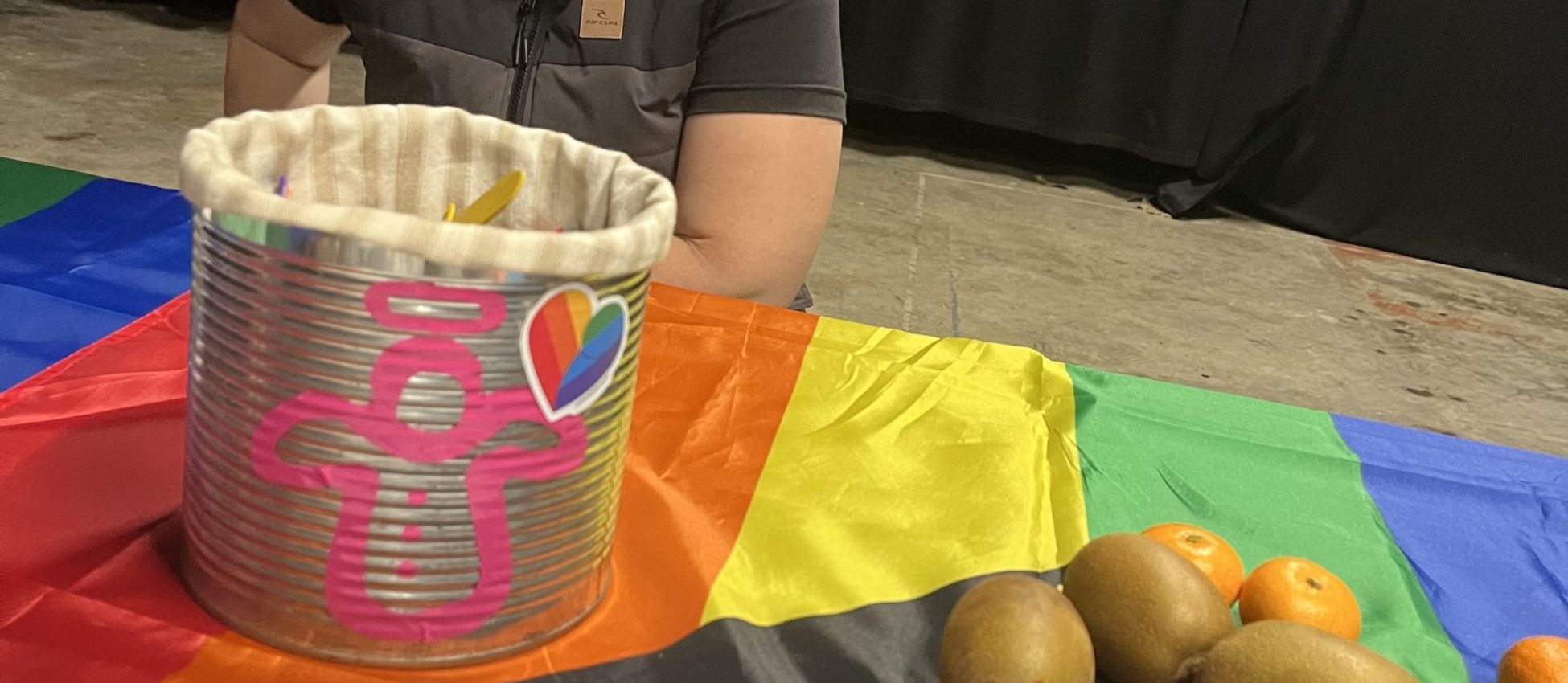 A person sits by a table draped in a rainbow flag